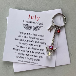 May include: A white card with the text "July Guardian Angel" and a poem about a guardian angel. The card is attached to a silver charm with a red crystal and a silver angel charm.