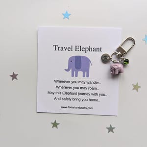 Könnte beinhalten: Ein lila Elefanten-Schlüsselanhänger mit einem silbernen Anhänger und einem grünen Kristall. Der Schlüsselanhänger wird mit einer Karte geliefert, auf der "Travel Elephant" steht, und einem Gedicht über sicheres Reisen.