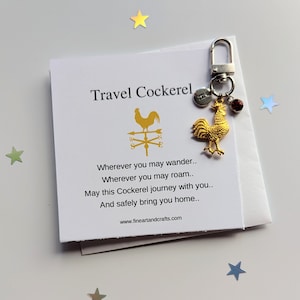 May include: A gold-coloured rooster keychain with a compass and a red gemstone charm. The keychain comes with a card that reads "Travel Cockerel" and a poem about safe travels.