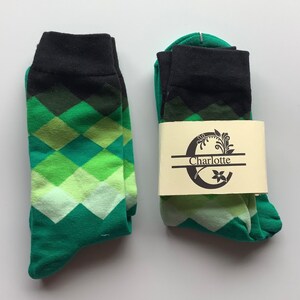 Puede incluir: Un par de calcetines verdes y negros con un patrón de diamantes. Los calcetines están empaquetados en una caja blanca con el nombre "Charlotte" impreso en ella.