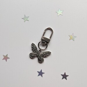 May include: A silver butterfly keychain with a silver metal ring and a silver clip. The keychain is on a white background with silver glitter stars.