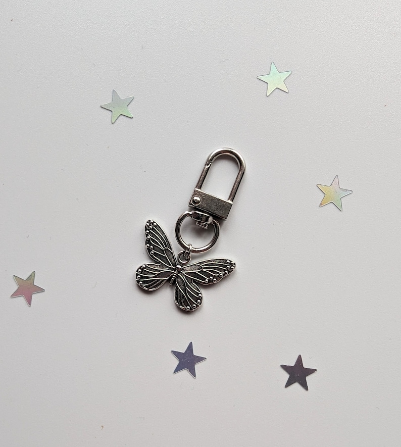 May include: A silver butterfly keychain with a silver metal ring and a silver clip. The keychain is on a white background with silver glitter stars.