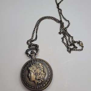 May include: Antique bronze-colored pocket watch with a chain. The watch face features a detailed relief of a woman's profile, the words "US UNUM", and the year "1877". The chain is also bronze-colored and has a clasp.