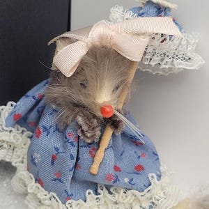 May include: A miniature figurine of a mouse wearing a blue floral dress with white lace trim and a matching bonnet. The mouse holds a small parasol and has a pink bow on its head. The mouse has gray fur and a red nose.