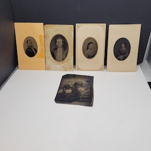 May include: A collection of five vintage photographs including four rectangular cards with oval portraits of individuals and one smaller, dark, rectangular tintype featuring two people sitting outdoors in a landscape setting.