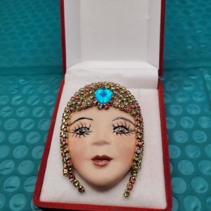 May include: A vintage brooch featuring a porcelain face with a blue rhinestone in the center, surrounded by a halo of colorful rhinestones. The brooch is in a red velvet box.