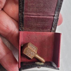 May include: A small brass teetotum spinning top with the text "TAKE ALL" and "PUT" engraved on its sides, resting inside a dark brown leather case with a red interior lining. The spinning top features a metallic finish and a short handle.