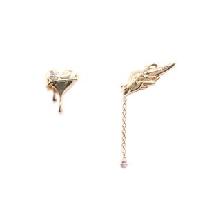 May include: A pair of gold-toned asymmetrical earrings. One earring is a heart with a dripping design, and the other is a wing with a chain and a dangling pink gem. The earrings are set against a white background.