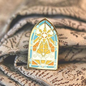 May include: A gold-toned metal enamel pin shaped like a pointed arch window. The design features a stained glass aesthetic with light blue, bright yellow, and orange sections depicting a sunburst, geometric lines, and stylized clouds.