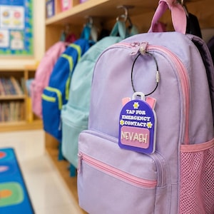 Puede incluir: Una etiqueta de mochila morada con forma de bolso pequeño con detalles de flores blancas y amarillas. La etiqueta presenta el texto blanco "TAP FOR EMERGENCY CONTACT" y una placa de nombre rosa con el nombre blanco "NEVAEH" en un lazo de alambre negro.