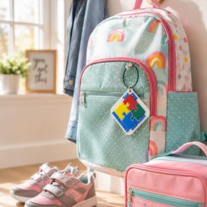 May include: A light blue backpack with a rainbow pattern and a white diamond-shaped tag attached to the front zipper. The tag features a four-piece puzzle design in red, yellow, green, and blue with the text TAP FOR EMERGENCY CONTACT.