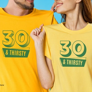 May include: Two yellow t-shirts with the text "30 & THIRSTY" in green. The number "30" has a 3D effect. The shirts are being held up by two people. The background is a solid blue.