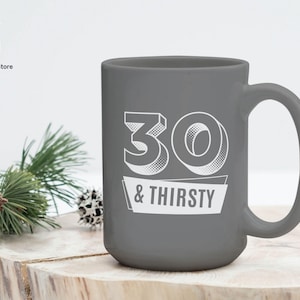 May include: A gray ceramic mug with the text "30 & THIRSTY" in white. The mug has a large handle and a simple, modern design. The mug is sitting on a wooden surface.