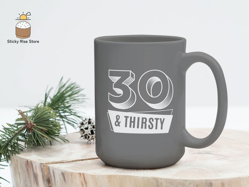 May include: A gray ceramic mug with the text "30 & THIRSTY" in white. The mug has a large handle and a simple, modern design. The mug is sitting on a wooden surface.