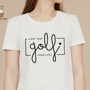 May include: White t-shirt with black text that reads "Livin' that golf Mom Life" with a small heart graphic.