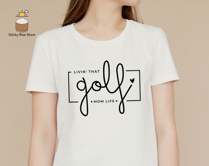 May include: White t-shirt with black text that reads "Livin' that golf Mom Life" with a small heart graphic.