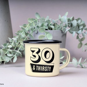 May include: Cream-colored enamel mug with a black rim and handle. The mug features the text "30 & THIRSTY" in a bold, black font. The mug is likely used for beverages and is a humorous gift for someone turning 30.