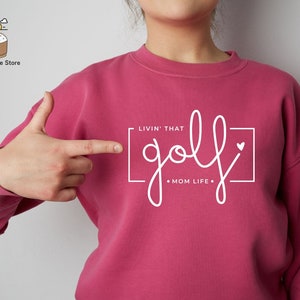 May include: Pink sweatshirt with white text that reads "Livin' That golf Mom Life" in a square frame with a small heart.