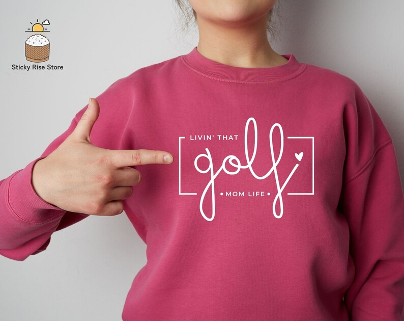 May include: Pink sweatshirt with white text that reads "Livin' That golf Mom Life" in a square frame with a small heart.