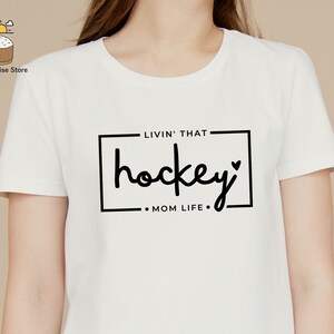 Może przedstawiać: Biała koszulka z czarnym napisem "Livin' that hockey Mom Life."