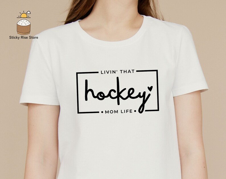 Może przedstawiać: Biała koszulka z czarnym napisem "Livin' that hockey Mom Life."