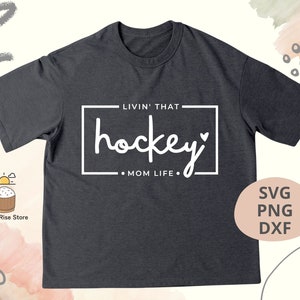 Może przedstawiać: Szary t-shirt z napisem "Livin' that hockey mom life." w kolorze białym. Tekst znajduje się w ramce z sercem na końcu słowa "hockey".