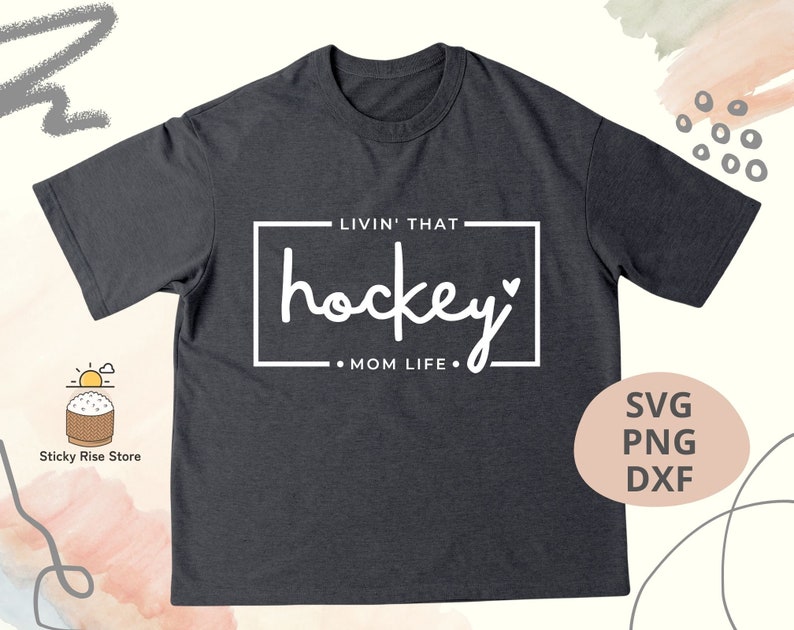 Może przedstawiać: Szary t-shirt z napisem "Livin' that hockey mom life." w kolorze białym. Tekst znajduje się w ramce z sercem na końcu słowa "hockey".