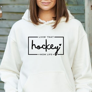 Może przedstawiać: Biała bluza z kapturem z czarnym napisem "Livin' that hockey mom life."
