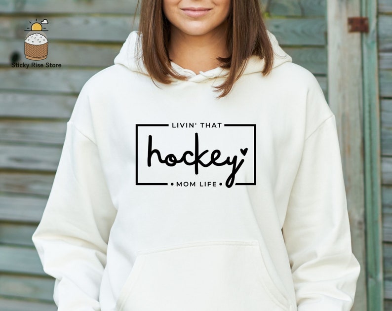 Może przedstawiać: Biała bluza z kapturem z czarnym napisem "Livin' that hockey mom life."