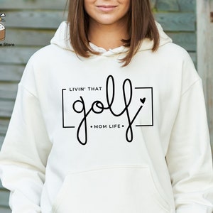May include: White hoodie with a black graphic that says "Livin' that golf - Mom Life" with a small heart.
