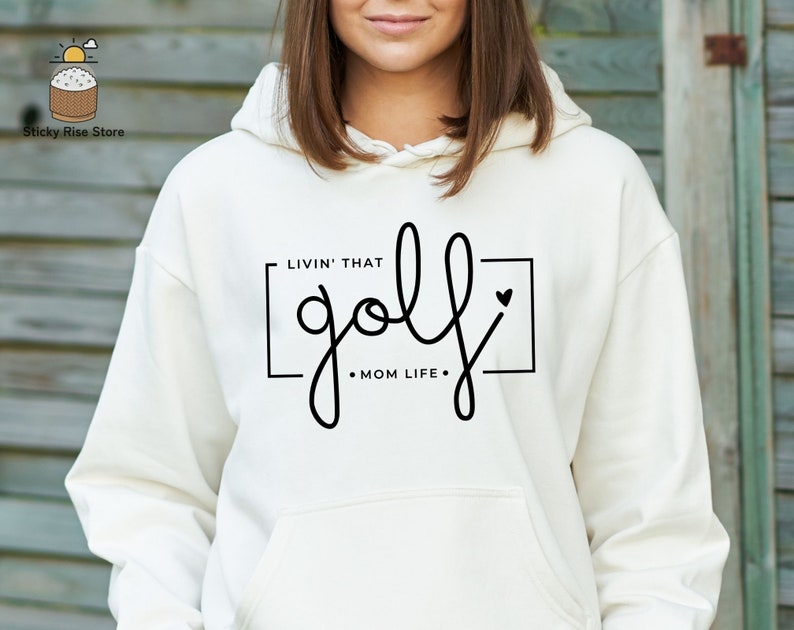 May include: White hoodie with a black graphic that says "Livin' that golf - Mom Life" with a small heart.