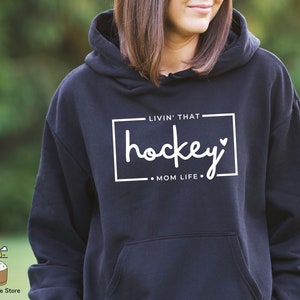 Może przedstawiać: Czarna bluza z kapturem z białym napisem "Livin' that hockey mom life."
