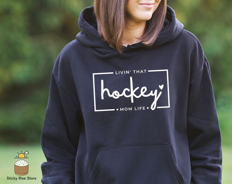 Może przedstawiać: Czarna bluza z kapturem z białym napisem "Livin' that hockey mom life."