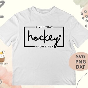 Może przedstawiać: Biała koszulka z czarnym napisem "Livin' that hockey mom life." Tekst znajduje się w ramce z symbolem serca.