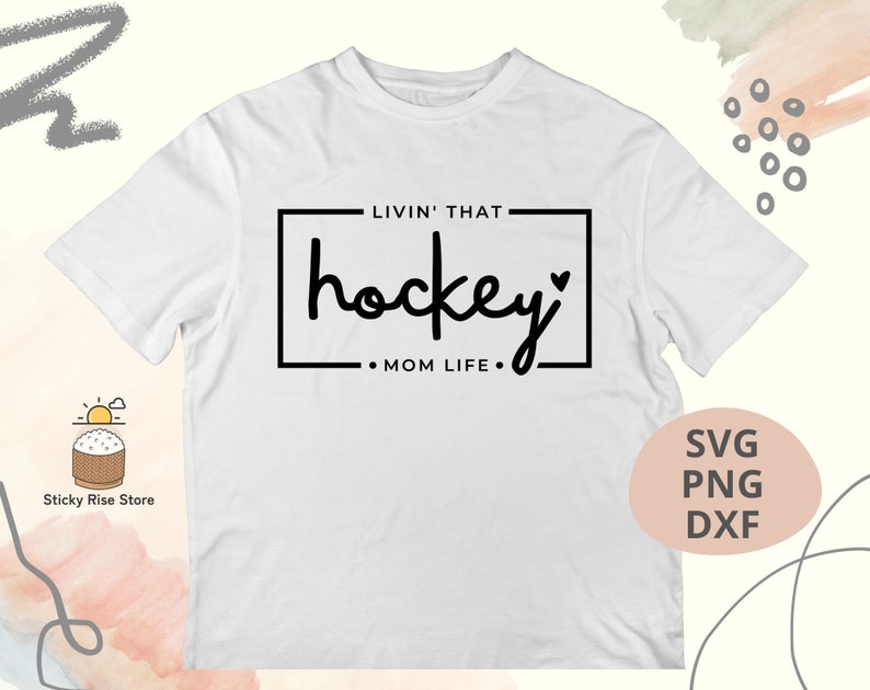 Może przedstawiać: Biała koszulka z czarnym napisem "Livin' that hockey mom life." Tekst znajduje się w ramce z symbolem serca.