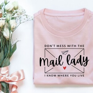 May include: A pink t-shirt with the text "Don't mess with the mail lady I know where you live" printed on it. The text is in a black font with a small red heart.