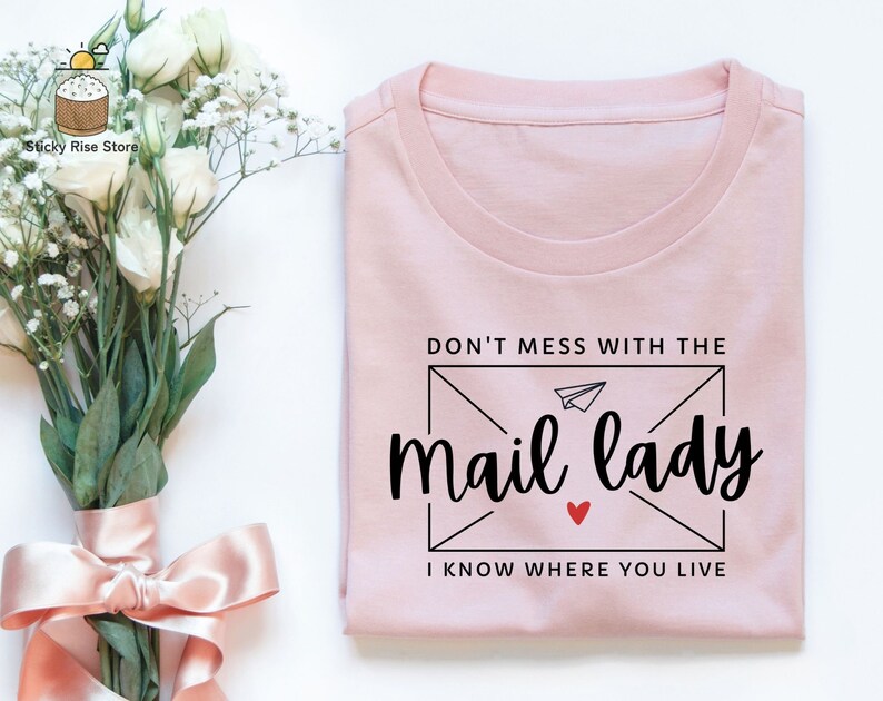 May include: A pink t-shirt with the text "Don't mess with the mail lady I know where you live" printed on it. The text is in a black font with a small red heart.