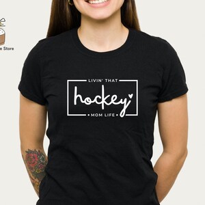 Może przedstawiać: Czarny T-shirt z białym tekstem, kt&oacute;ry brzmi "Livin' that hockey mom life" w białym prostokącie.