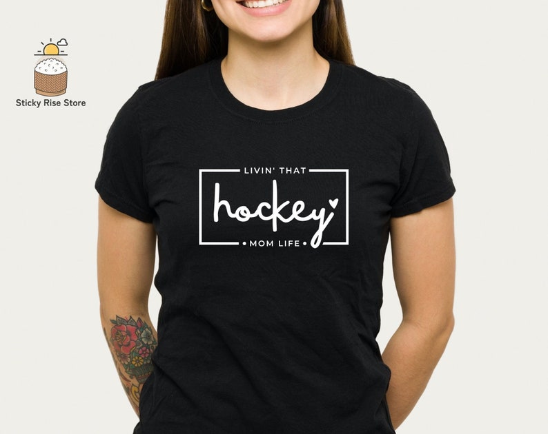 Może przedstawiać: Czarny T-shirt z białym tekstem, kt&oacute;ry brzmi "Livin' that hockey mom life" w białym prostokącie.