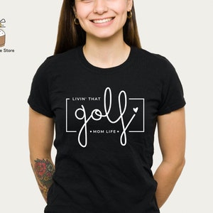 May include: Black t-shirt with white text that says "Livin' that golf mom life."  The text is inside a white rectangle with a small white heart in the top right corner.