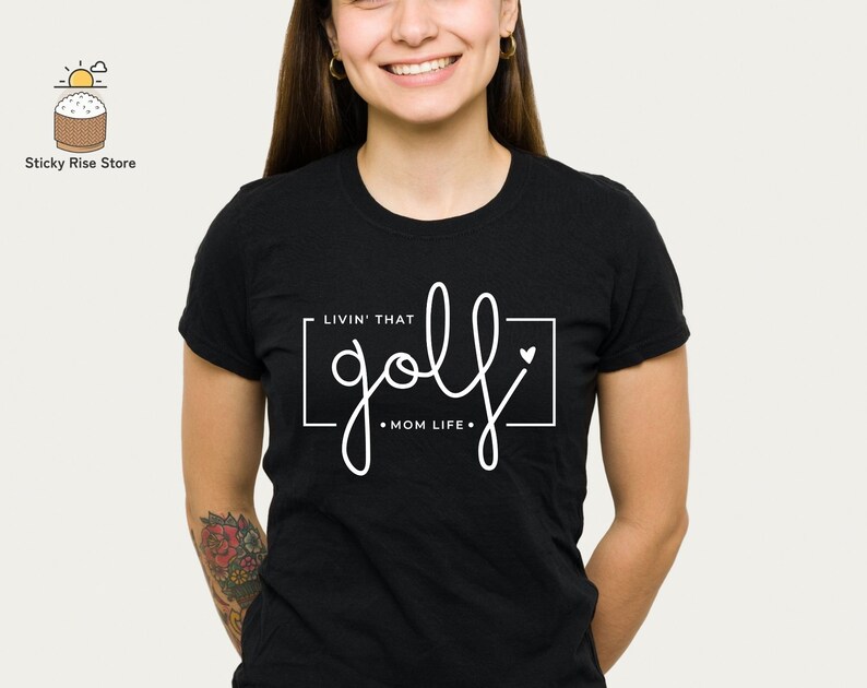 May include: Black t-shirt with white text that says "Livin' that golf mom life."  The text is inside a white rectangle with a small white heart in the top right corner.
