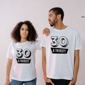 May include: Two people wearing white t-shirts with the text "30 & THIRSTY" in black. The number "30" is in a bold, outlined font above the words "& THIRSTY" on a black banner. The shirts are short-sleeved.