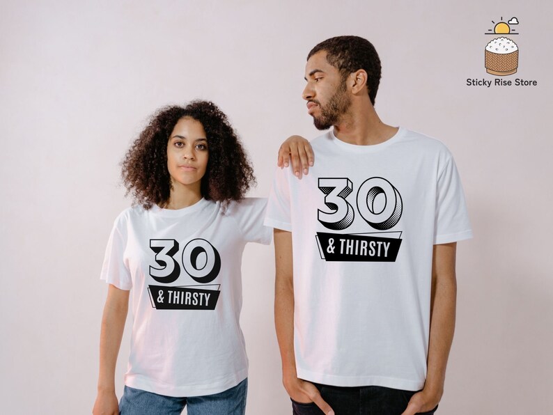 May include: Two people wearing white t-shirts with the text "30 & THIRSTY" in black. The number "30" is in a bold, outlined font above the words "& THIRSTY" on a black banner. The shirts are short-sleeved.