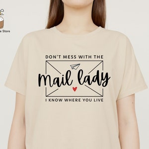 May include: Beige t-shirt with a black graphic that says "Don't mess with the mail lady I know where you live" with a red heart and a paper airplane inside an envelope.