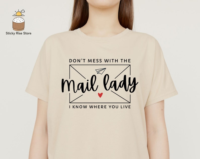 May include: Beige t-shirt with a black graphic that says "Don't mess with the mail lady I know where you live" with a red heart and a paper airplane inside an envelope.