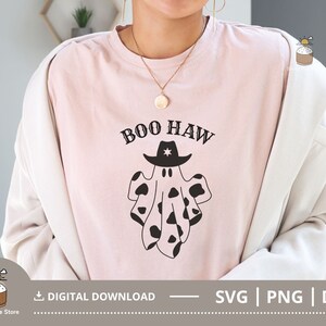 Puede incluir: Una camiseta rosa con un fantasma blanco y negro que lleva un sombrero de vaquero. El fantasma tiene un patr&oacute;n de vaca. El texto "BOO HAW" est&aacute; impreso sobre el fantasma.