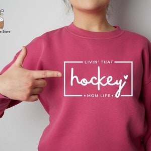 Może przedstawiać: R&oacute;żowa bluza z nadrukiem "Livin' That hockey Mom Life" z przodu.
