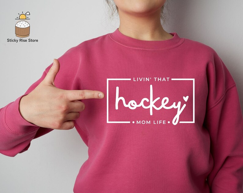 Może przedstawiać: R&oacute;żowa bluza z nadrukiem "Livin' That hockey Mom Life" z przodu.