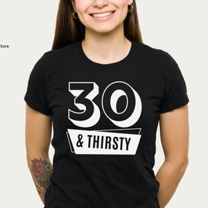 May include: Black crew neck t-shirt with the text "30 & THIRSTY" in white block letters. The number 30 is large and bold, with the words "& THIRSTY" on a white banner below. The shirt is worn by a person.