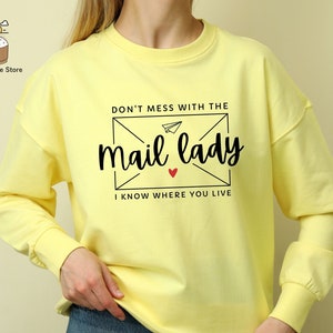 May include: Yellow long-sleeve sweatshirt with a black graphic print of an envelope with a paper airplane and the text "Don't mess with the mail lady I know where you live".